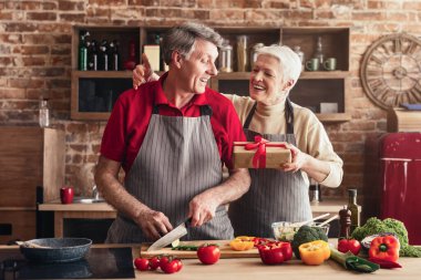 Mutfakta kıdemli bir çift birlikte yemek hazırlıyor. Adam sebzeleri doğrarken kadın elinde bir hediye tutuyor ve ona gülümsüyor..