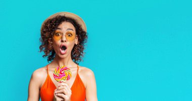 Elinde renkli bir lolipop tutan turuncu mayo giymiş genç siyah bir kadın ve arkasında mavi bir arka planla şaşırmış görünüyor. Ağzı açık, güneş gözlüğü ve hasır şapka takıyor..