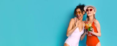 Mayo giymiş, güneş gözlüğü takan ve gülümseyen iki kadın mavi arka planda kokteyl içerken. Yan yana duruyorlar ve arkadaş gibi görünüyorlar. Kumsalda ya da havuzda eğleniyorlar..