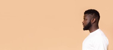 Kısa saçlı ve sakallı Afro-Amerikan bir adam. Beyaz bir tişört giyiyor ve şeftali arkaplanının önünde duruyor. Fotokopi alanı olan bir panorama.