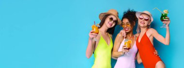 Three multiracial girlfriends wearing swimsuits and sunglasses, laughing and holding colorful cocktails against a blue background. They appear to be having a fun and carefree summer day, copy space clipart