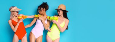 Mavi arka planda üç genç kadın mayolarıyla poz veriyor. Ellerinde su tabancaları var ve neşeli görünüyorlar. Yaz havuzu partisinde ya da plajda eğleniyor gibi görünüyorlar..