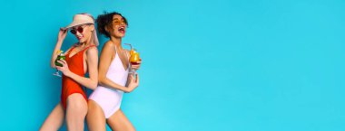 İki tane çok ırklı mayo giymiş genç kadın parlak mavi bir arka planın önünde duruyor. İkisinde de kokteyller var ve gülümsüyorlar.