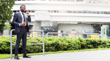 Yakışıklı afro-amerikan işadamı kahve molası sırasında cep telefonu kullanıyor, korkulukların önünde duruyor. Tam uzunluk
