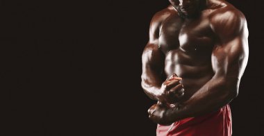 Kaslı bir adamın kaslarını esnetirken, fiziksel özelliklerini sergilediği yakın plan bir fotoğraf. Kırmızı şort giyiyor ve siyah arka planda duruyor..