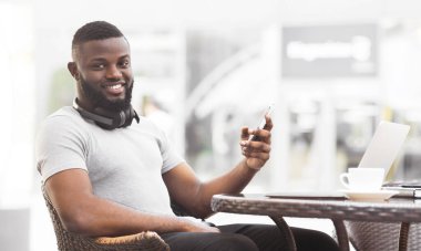 Kafe masasında dizüstü bilgisayarı ve bir fincan kahveyle oturan genç bir adam akıllı telefonuna bakıp gülümsüyor..