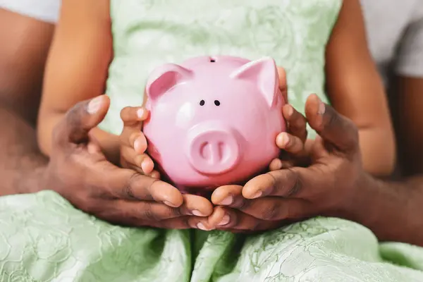 stock image A child and parent are holding a pink piggy bank. The child is wearing a green dress and the parent is wearing a white shirt. The piggy bank is in the center of the image, cropped