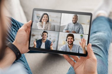 Adam elinde bir tablet tutuyor ve dört kişiyle online video konferansına katılıyor, evdeki kanepede dinleniyor.