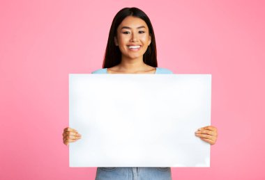 Uzun siyah saçlı Asyalı genç bir kadın pembe bir arka planın önünde duruyor. Önünde büyük, boş beyaz bir tabela tutuyor..