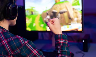 Ekose gömlekli bir oyuncu bilgisayarında video oyunu oynarken zaferi kutlar. Kulaklık takıyorlar ve yumruklarını havaya kaldırıyorlar..