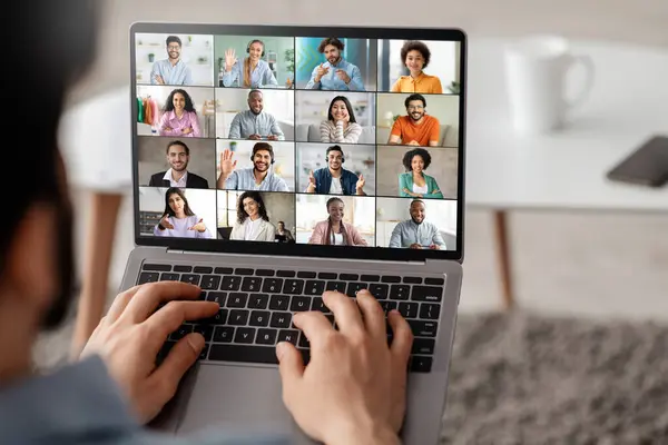 stock image A man is sitting on a couch and using a laptop to participate in a video conference. The screen of the laptop is displaying a grid of video feeds showing several different people