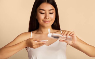 Uzun siyah saçlı genç bir Asyalı kadının pamuk pede yüz toneri uyguladığı görülüyor. Gülümsüyor ve elindeki pamuğa bakıyor..