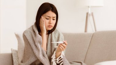 Battaniyeye sarılı bir kadın kanepeye oturur ve ateşini termometreyle ölçer. Endişeli görünüyor..