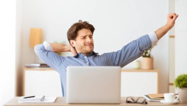 Bir adam evindeki bir masada oturur, kollarını ve sırtını uzatır. Biraz yorgun görünüyor..