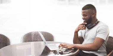 Bir adam dışarıda bir kafede oturuyor ve laptopuyla çalışıyor. Boynunda kulaklık var ve gülümsüyor..