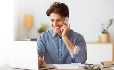 Bir adam dizüstü bilgisayarla çalışırken gülümsüyor ve telefonla konuşuyor. Bir masada oturuyor ve önünde evrak işleri var..