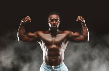 Kaslı bir adam kaslarını esnetir, gücünü ve fiziğini ince duman efektleriyle karanlık bir arka plana karşı sergiler..