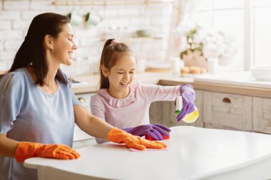 Bir anne ve kızı mutfak masalarını birlikte temizliyorlar. Anne turuncu eldiven giyip masayı bezle silerken, kız mor eldiven giyip sprey sıkar.