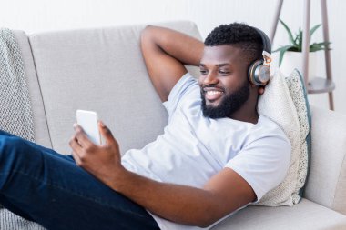 Sakallı genç siyah bir adam evdeki koltukta dinleniyor, kulaklık takıyor ve akıllı telefonunu kullanıyor. Gülümsüyor ve eğleniyor gibi görünüyor..