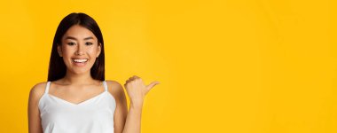 Uzun siyah saçlı Asyalı genç kadın gülümsüyor ve fotokopi alanını işaret ediyor. Beyaz bir atlet giyiyor ve parlak, dostane bir ifadesi var. Arka plan koyu sarı renkte, panorama.