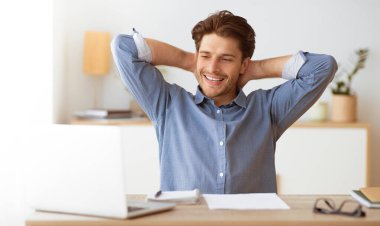 Bir adam masasında oturuyor, gülümsüyor ve elleri başının arkasında, dizüstü bilgisayara bakarken rahatlıyor..
