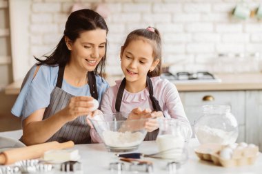 Bir anne ve küçük kızı mutfakta birlikte yemek pişiriyorlar. Anne bir yumurta tutuyor ve gülümsüyor, kız ise bir kase una bakıyor. İkisi de önlük takıyor.