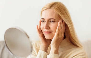 Nazikçe gülümseyen sarışın bir kadın aynada tenini kontrol eder, ellerini yanaklarına tutar..