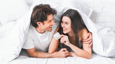 Bir çift yatakta uzanıyor, ikisi de gülümsüyor, elinde prezervatif tutan kadınla. Örtünün altındalar ama başları ve omuzları görünüyor..