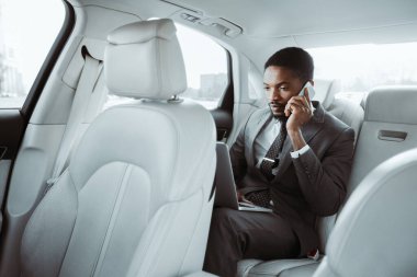 Afrikalı Amerikalı iş adamı bir arabanın arka koltuğunda oturuyor, dizüstü bilgisayarla çalışıyor ve telefonla konuşuyor. İşine odaklanırken ciddi bir konuşma yapıyor gibi görünüyor..