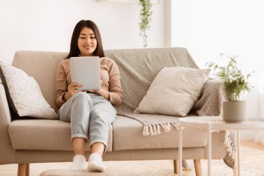Oturma odasındaki kanepede oturan bir kadın, rahatlamış ve tablet üzerinde geziniyor..