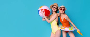 Bu resim, mayo ve hasır şapka giyen iki gülümseyen kadını gösteriyor. Mavi arka planda bir plaj topu ve şişme bir şamandıra tutuyorlar. Görüntü yaz eğlencesini ve tatilini çağrıştırıyor..