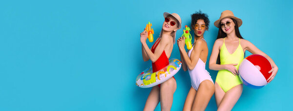 Three women are posing in swimsuits with water guns and a beach ball on a bright blue background. They appear to be having fun and enjoying the summer, copy space