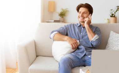 Mavi gömlekli bir adam beyaz bir koltukta oturuyor, telefonda konuşurken gülümsüyor..