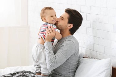 Bir adam yatak odasında bir yatakta oturuyor. Kollarında bir bebek tutuyor ve bebeği yanağından öpüyor. Bebek gülümsüyor ve mutlu görünüyor.