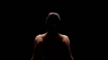 Sırtı kameraya dönük oturan bir kadın tek bir ışık kaynağıyla aydınlatılır. Yüzünü izleyiciden ayırıyor ve arka plan tamamen siyah..