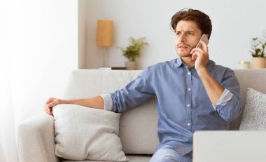 Bir adam kanepede oturur, gözlerini kaçırırken cep telefonuyla konuşur. Mavi bir gömlek giyiyor ve önünde bir laptop var..