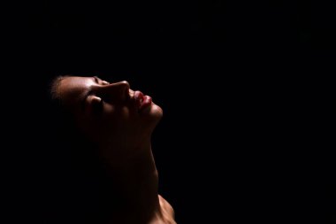 Tek bir ışık kaynağı tarafından aydınlatılmış, gözleri kapalı ve dudakları hafifçe ayrılmış bir kadının yakın çekim görüntüsü. Arkaplan tamamen siyah, kopyala alanı