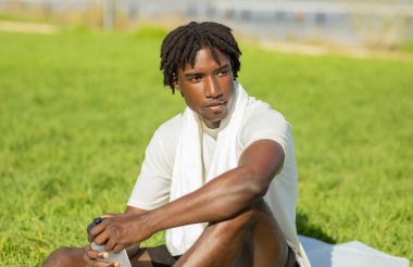 Rastalı Afrikalı Amerikalı bir adam çimenli bir tarlada oturuyor, beyaz bir tişört giyiyor ve boynunda beyaz bir havlu var. Elinde bir su şişesi var ve idmandan sonra dinleniyor gibi görünüyor..