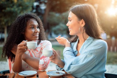 Bu görüntü, güneşli bir öğleden sonra dışarıda bir masada oturmuş kahvelerini yudumlayan iki kadını gösteriyor. Yakın arkadaş gibi görünüyorlar, gülüyorlar ve sohbet ediyorlar. Sahne rahat ve rahat.