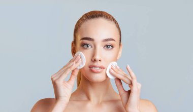 Bu görüntü, yüzüne pamuk mermileri süren tarafsız bir yüzü olan genç bir kadını gösteriyor. Bir stüdyo ortamında, açık mavi bir arka planın önünde duruyor..