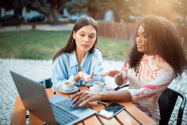 İki kadın bir kafenin dışında bir masada oturuyorlar. Bir kadın dizüstü bilgisayar kullanırken diğer kadın el kol hareketi yapıp bakıyor. Bilgisayarın yanındaki masada bir cep telefonu var..