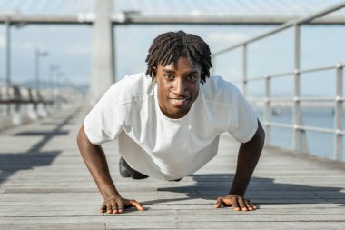 Afrika kökenli, rastalı bir adam, okyanusun kıyısındaki ahşap bir güvertede şınav çekiyor. Beyaz bir tişört giyiyor ve iyi durumda gibi görünüyor. Gülümsüyor ve kameraya bakıyor.