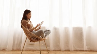 Genç bir kadın pencerenin yanındaki hasır sandalyede oturuyor, tabletini karıştırıyor..