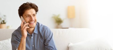 Kanepede oturan bir adam telefonla konuşuyor, gülümsüyor ve soluna bakıyor..