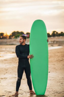 A man wearing a black wetsuit stands on a sandy beach holding a green surfboard. The sun is setting in the background, casting a warm glow over the scene. clipart