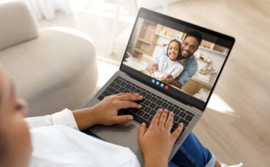 Siyah kadın anne kanepeye oturur ve ailesiyle görüntülü konuşma için dizüstü bilgisayar kullanır. Dizüstü bilgisayardaki adam ve genç bir kız, ikisi de gülümsüyor ve mutlu görünüyorlar..