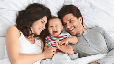 Bu resim üç kişilik mutlu bir aileyi gün boyunca yatakta gösteriyor. Ebeveynler gülümsüyor ve babalarının yüzüne dokunmak için gülümseyip uzanan bebeklerine bakıyor..