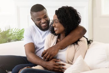 Mutlu bir çift beyaz bir kanepede oturur, adam kolunu kadının omzuna atar ve ikisi de gülümserler..