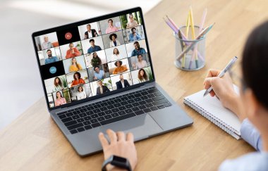 Kadın yönetici, önlerinde dizüstü bilgisayarla ahşap bir masada oturuyor. Ekranda birden fazla katılımcının katıldığı bir video konferansı gösteriliyor.