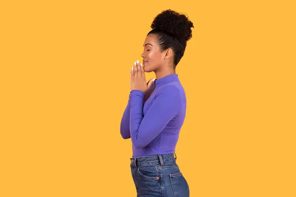 stock image A young woman with her hair in a bun stands in profile with her eyes closed and hands clasped together in prayer. She is wearing a purple long-sleeve shirt and blue jeans.